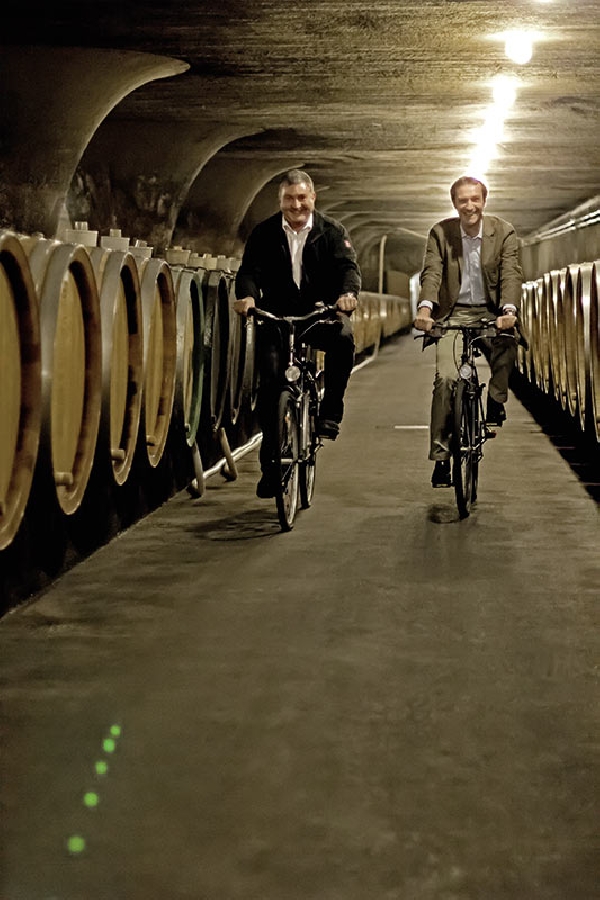 Das Foto zeigt Karsten Weyand und Johannes Becker bei einer Fahrt auf dem Rad durch den Weinkeller.